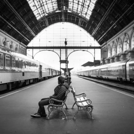 Osoba siedząca na ławce na stacji kolejowej, w tle pociągi i architektura budynku w stylu secesyjnym.
